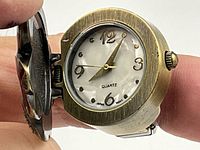 Close-up image of the round quartz watch face under the flip-up cover of the ring watch with gold-tone numbers and mother-of-pearl dial.