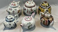 Top front view of the collection of 6 painted lidded tea pots showing various decoration styles and colors