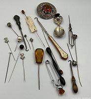 Collection of approximately 20 hat pins and related items shown laid out on white surface, measurement reference included