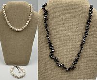 White pearl choker necklace displayed on bust with matching bracelet at base; partial view of black pearl necklace beside.