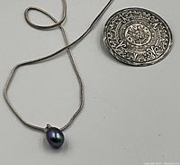 Necklace with black pearl pendant displayed next to a circular sterling silver brooch with detailed design for size reference.