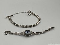 Two silver bracelets displayed side by side on white background, showing the spherical silver bead bracelet with toggle clasp and the silver bracelet with an oval Delft ceramic centerpiece, accompanied by a green apple for size reference.