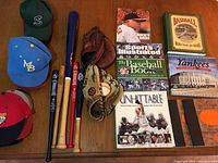 Photo showing several baseball caps, three wooden baseball bats, two baseball gloves, and multiple baseball-themed books including Sports Illustrated, The Baseball Book, and Unforgettable.