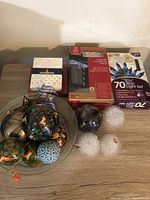 Christmas ornaments and outdoor light sets displayed on a wood surface, including spherical hand-painted ornaments in a glass bowl, snowflake shaped lights, and packaged blue LED light set.