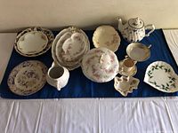 First photo shows an overview of the various dishes arranged on a dark blue cloth. Includes plates, pitcher, teapot, covered serving bowl, cups, and small decorative dishes.