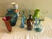 Front view of 11 assorted glass and ceramic vases including tall porcelain, red glass trumpet-shaped, blue ribbed glass, and various round-bottomed glass vases in multiple colors on a white table.
