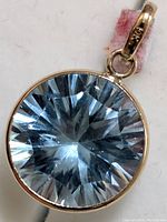 Close-up front view of round faceted natural blue topaz set in 10K gold bezel pendant with bail, showing gemstone clarity and faceting details.