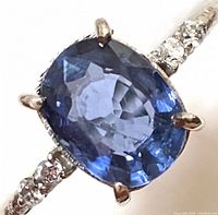 Close-up image showing cushion-cut blue sapphire with prong setting and small lab diamond accents on gold band.