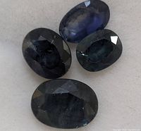 Four oval faceted dark blue sapphires arranged on a plain surface, showing color and facet structure.