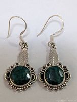 Close-up of a pair of silver earrings with green faceted round emerald stones, decorative silver details including leaf motifs, and hook-style ear wires.