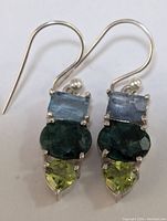 Photo of pair of silver earrings with three gemstones each: rectangular blue, oval emerald, heart-shaped peridot.
