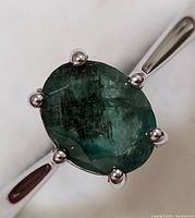Close-up image of silver ring showing oval green emerald gemstone held by six prongs.