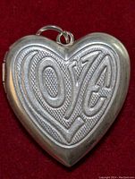Front view of a silver heart-shaped locket pendant featuring embossed stylized text 'LOVE' with textured background, showing the pendant's loop bail.