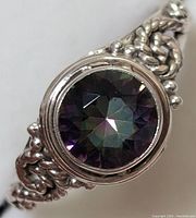 Close-up image of a silver ring featuring a round faceted mystic topaz gemstone with braided and beaded silver band details.