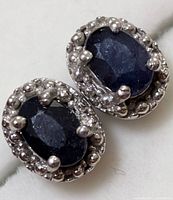 Close-up of two silver sapphire earrings showing oval dark blue sapphire stones surrounded by small clear rhinestones in detailed silver settings.