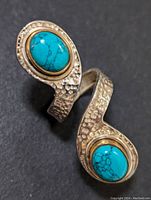 Silver ring shown on black background. Ring has two turquoise stones set on open wrap-around V-shaped silver band with hammered texture.