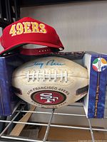San Francisco 49ers red hat positioned behind football showing 49ERS embroidery and football with signature and logo in box.