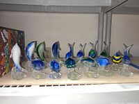 Full collection of glass dolphin and fish figurines displayed on a shelf showing 12 unique pieces in blue, white, green, and yellow colors, each on clear glass bases.