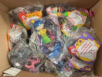 Photo showing box filled with mostly unopened McDonald's Happy Meal animal and character toys in original sealed plastic packaging.