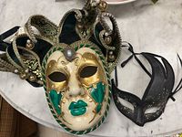 Top-down view of the two masks showing the green and gold jester mask with velvet and pearl details and the black glitter mask.