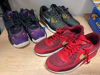 Three pairs of men's Nike sneakers displayed on wooden surface. Black/purple pair with red Nike swoosh and Velcro strap, green/yellow Nike Air Max pair, and red/gold pair with gold swoosh. Shoes show signs of use with dirt on soles.