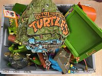 Top view of gray bin filled with assorted TMNT figures and vehicles, including green shell, orange half shell, and green tubular.