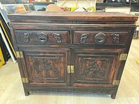 Front view of dark wooden cupboard with Asian detailed carvings on drawers and doors and brass hinges.