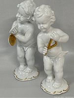 Pair of Schaubach Kunst porcelain cherubs standing side by side, showing front and side views with detailed gold accents on instruments and base.