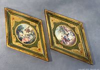Pair of diamond-shaped Florentine wood wall hangings shown side by side on gray surface, displaying ornate carving, green and gold paint, and central oval romantic scenes.