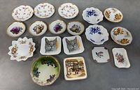 Top-down view of 19 miniature porcelain plates with varied floral, animal, and scenic designs arranged in rows on flat surface.