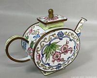 Side view of the oval enameled mini teapot showing pink and blue floral patterns with gold scrollwork, square lid with brown knob, and brown handle.