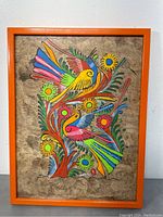 Full frontal image of the framed painting showing vibrant birds and flowers on brown amate paper with an orange frame.