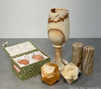 Photo shows stone goblet, two cylindrical salt and pepper shakers, boxed set of two spherical salt and pepper shakers, and two geometric multifaceted stone objects on gray surface with white background.