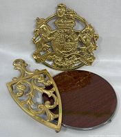 Three trivets including ornate Kings Arms brass trivet, shield-shaped brass trivet, and round Formica trivet with chrome rim and label.