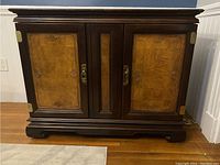 Closed view of Asian style vintage bar server cabinet with carved mahogany and ash burl panels on doors. Brass hardware visible. Cabinet sits on floor against a blue wall.