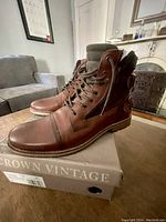 Pair of Crown Vintage men's brown leather boots shown on top of a Crown Vintage shoebox in a well-lit room, showcasing the profile and design details.