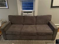 Front view of gray three-seater couch showing three seat and three back cushions, firm armrests, placed in a room with white window and gray carpet.