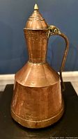 Front angle photo of the copper watering jug showing the wide base, tapered body, brass handle, and pointed lid with finial.