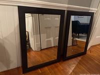 Two black framed rectangular mirrors side by side leaning against a wall, reflecting hardwood floor and part of a doorway.
