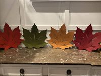 Four autumn leaf-shaped placemats in red, green, orange, and burgundy placed side by side on a brown marble countertop against a white paneled wall.