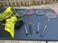 Four badminton rackets laid on dark surface alongside badminton net and bag