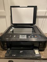 Front angled open view showing flatbed scanner glass and control panel with buttons and small display. Printer setup on marble surface.