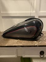 Side view of Honda motorcycle gas tank, showing black and gray coloring with red trim and Honda wing logo.