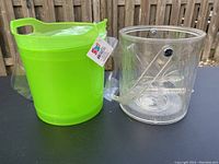 Two ice buckets placed next to each other outdoors on a black surface in front of a wooden fence. Green bucket with handle and retail tag is on the left. Clear bucket with handle and etched design is on the right.