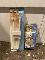 Photo showing both the 6-inch cabinet slide lock and the 4-pack ultra secure drawer locks in original packaging, white plastic locks by Safety 1st