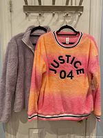 Photo of two girls' sweaters hanging on a rack: one fuzzy light purple zip-up jacket and one colorful Justice pullover with stripes and "JUSTICE 04" text.