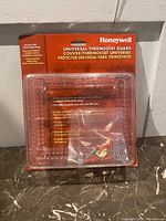 Front view of sealed Honeywell Universal Thermostat Guard packaging showing product inside hard plastic blister pack with mounting hardware and red and white cardboard backing.