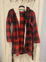 Full view of kids robe hanging showing red and black plaid pattern, hood, pockets, and open front.