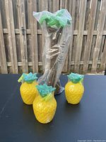 Photo showing three yellow pineapple-shaped plastic beverage cups and one tall palm tree-shaped cup in the background, all with plastic straws, displayed on a black surface in front of a wooden fence.