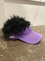 Side angle of purple hat with attached black faux spiky hairpiece placed on a brown marble surface.